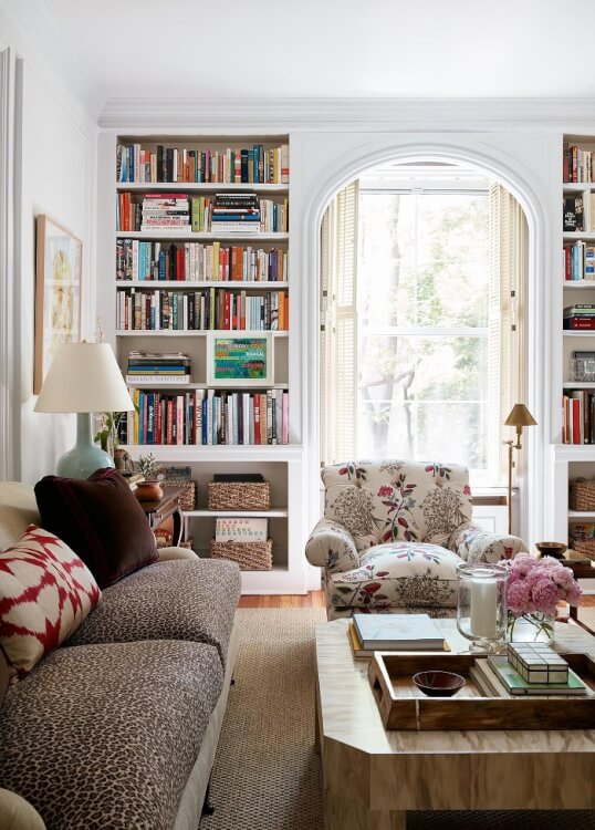 Hình ảnh phòng khách với sofa xám, tối tựa màu sắc, bàn trà kính, ghế bành họa tiết hoa lá, cạnh đó là kệ sách kịch trần