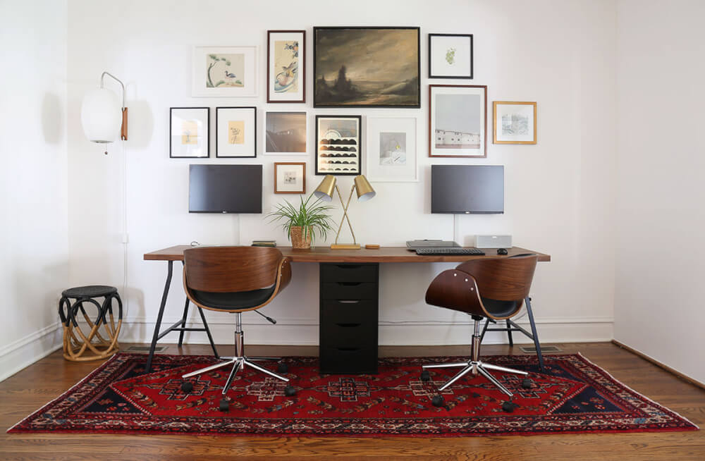 Hình ảnh toàn cảnh văn phòng tại nhà dành cho 2 người với bộ bàn ghế đóng sẵn, thảm trải thổ cẩm tông màu đỏ và bộ tranh treo tường bắt mắt