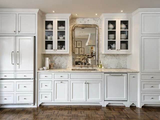 hình ảnh phòng bếp với gương khung bạc treo trên bồn rửa, xung quanh là hệ tủ kệ lưu trữ màu trắng chủ đạo