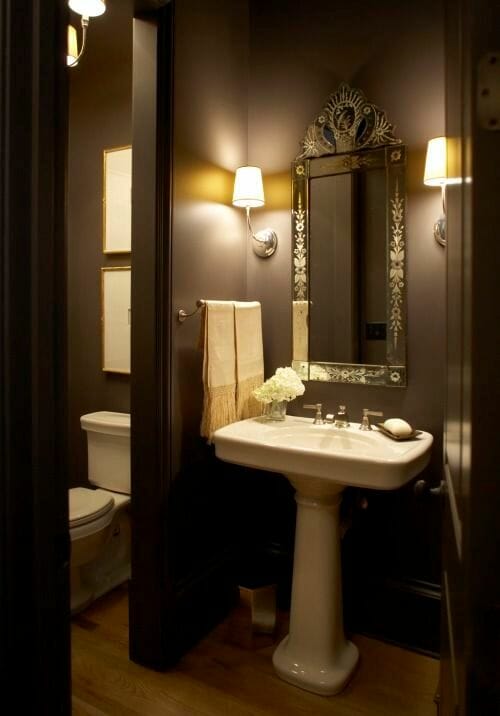 hình ảnh phòng tắm màu nâu sành điệu với gương soi kiểu dáng cổ điển, bồn rửa đứng và đèn tường ánh sáng vàng ấm áp.