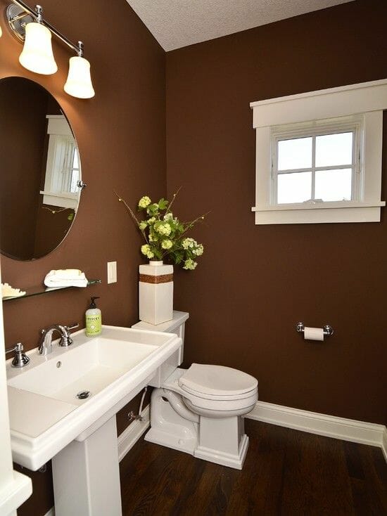 Hình ảnh phòng tắm với tường sơn màu nâu sô cô la, sàn lát gỗ cùng tông màu, gương lớn gắn tường, đèn màu trắng