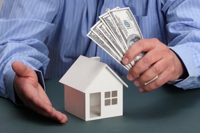 hình ảnh một người mặc sơ mi màu xanh lam kẻ sọc nhỏ cầm tiền đô la, trên bàn là mô hình ngôi nhà