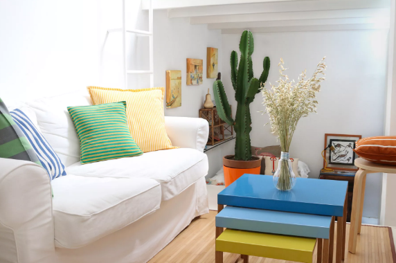Hình ảnh phòng khách với ghế sofa màu trắng, gối tựa sắc màu, bàn cà phê 3 lớp màu sắc, chậu cây xương rồng lớn